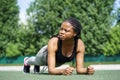 Determinated black woman does plank exercise on green turf flooring of city stadium against park