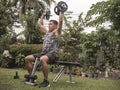 A fit and athletic middle aged asian guy does seated dumbbell shoulder presses on a bench at his front yard.