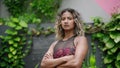 Confident multiracial woman in sportswear poses with crossed arms among green plants. Fit biethnic female model