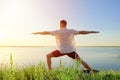 Fit muscular man doing yoga by the water on the grass with hands stretch. Orange sunlight sunset tranquility at lake Royalty Free Stock Photo