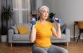 Fit senior woman working out with dumbbells, doing domestic training, exercising her biceps at home, sitting on fitball Royalty Free Stock Photo