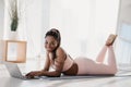 Fit young black lady using laptop computer, lying on sports mat, having break from domestic training, indoors Royalty Free Stock Photo