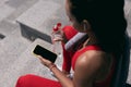 Fit young woman with dark hair in red sportswear and white earphones holds smartphone and water bottle. High angle cut