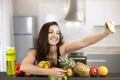 Fit young woman wearing sexy black sports top shooting selfie on her smartphone standing in the kitchen full of fruits, dietology