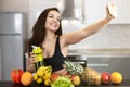 Fit young woman wearing sexy black sports top shooting selfie standing in the kitchen full of fruits, dietology and nutrition