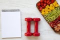 Fitness concept, diet plan. Dumbbells , notebook, chopped fruits on white wooden background,