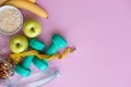 Fitness concept with dumbbells, nuts, bananas, apples and skipping rope on pink background with copy space.