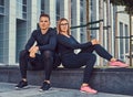 Fitness couple in a sportswear, blonde girl and a handsome muscular guy sitting on a bench against a skyscraper. Royalty Free Stock Photo