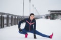 Fitness girl model runner doing flexibility exercise for legs before run at snow winter promenade Royalty Free Stock Photo