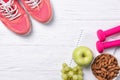 Fitness and healthy eating concept, pink sneakers and dumbbells with apple, grapes and almond nuts on notepad, wooden background