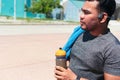 Fitness man uses water bottle while standing in park. Cheerful sportsman taking a break in a park