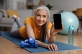 Fitness mobile app. Happy senior woman showing smartphone with blank screen, exercising at home, mockup Royalty Free Stock Photo