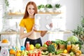 Fitness redhead woman with weight scale standing behind table with fruit and vegetables in kitchen