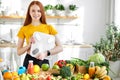 Fitness redhead woman with weight scale standing behind table with fruit and vegetables in kitchen