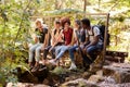 Five millennial friends sitting on a bridge in a forest talking during a hike, full length