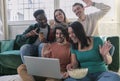 Five multi-ethnic millennial friends sitting on sofa looking at computer screen wave hands laughing enjoy distant communication