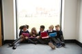 Five primary school kids sitting in a row on the floor in front of a window in a school corridor looking at tablet computers, fron