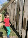 A five-year-old cute curly-haired black-haired boy in green pants and a red T-shirt looks like a painted wooden fence. The concept Royalty Free Stock Photo