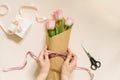 Flat female hands tie a satin ribbon bow on a simple bouquet of fresh pink tulip flowers in craft paper on a beige table