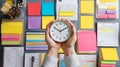 Flat lay composition showing hands holding a modern clock above a cluttered workspace filled with vibrant post-it notes, weekly Royalty Free Stock Photo