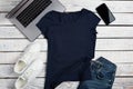 Flat lay of dark blue t-shirt lying on rough wooden planks with laptop, smartphone and apparel accessories.