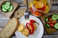 Flat lay of marinated cheese in olive oil with capsicum in glass jar, fork, plate tomato bread and cucumber on wooden background