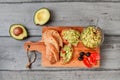 Flat lay photo, freshly prepared guacamole in small glass bowl, bread, tomatoes, olives at working board and two avocados next