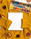 Flat lay of sketchbook, paints with brushes, yellow sweater, leaves, cinnamon sticks and pine cones on a white wooden background. Royalty Free Stock Photo