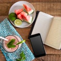 A flat lay square ratio breakfast tabletop photo with watermelon smoothie phone, book and sliced fruit on plate