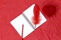 FLAT LAY STYLE. On a red crumpled paper surface there is a white notebook on springs and red pen, a feather of a bird and a heart