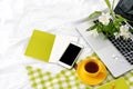 Flat lay tablet, phone, yellow cup of tea, laptop and flowers on white blanket with green napkin