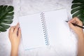 Flat lay of woman hand writing in a blank white page notebook on white marble desk, top view. green palm leaf on desk