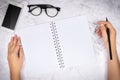 Flat lay of woman hand writing in a blank white page notebook on white marble desk, top view. glasses and mobile smart phone