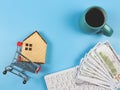 Flat layout of wooden house model in shopping trolley, computer keyboard, banknotes and blue cup of black coffee on blue Royalty Free Stock Photo