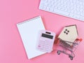 Flat layout of wooden house model in shopping trolley, pink calculator, blank page notebook  and computer keyboard  on pink Royalty Free Stock Photo