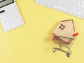 flat layout of wooden house model in shopping trolley, white calculator and computer keyboard on yellow background with copy Royalty Free Stock Photo