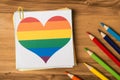 Flatlay close up photo of pile of paper stickers with drawn rainbow heart and multicolored pencils isolated wooden backdrop