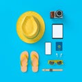 Flatlay with hat with sun glasses, toy plane, slippers, hat, suntan cream, phone and camera on blue minimal style background.