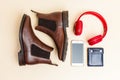 Flatlay of Pair of Classic Leather Chealsea Boots, Wireless headphones, Leather Wallet, Cellphone Placed Near One Another On Beige