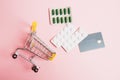 Flatlay, shopping cart, bank card and pills in a blister on a pink background