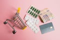 Flatlay, shopping cart, bank card and pills in a blister on a pink background