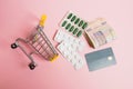 Flatlay, shopping cart, bank card and pills in a blister on a pink background