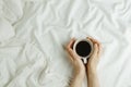 Flatlay of woman`s hands holding cup of coffee in bed on white sheets