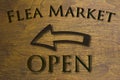 Flea Market Open sign set on a wood background and a left facing arrow. Brown 3D text against wood grain background.