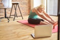 Flexible mature blonde woman in sportswear sitting on the floor, stretching her body while watching online video yoga Royalty Free Stock Photo