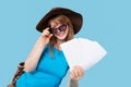 A flirtatious girl tourist in a hat with sunglasses poses and shows white empty space air ticket and flying documents to the