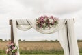 Floral wooden arch with white cloth and fresh violet pink white flowers with green leaves on a rustic wedding ceremony.