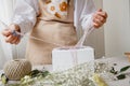 A florist decorates a gift box with flowers and a ribbon on a white desktop. Only the hands are in the frame