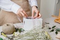 A florist decorates a gift box with flowers and a ribbon on a white desktop. Only the hands are in the frame