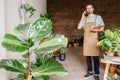 Floristry business and people concept. Worker in uniform carrying flower in pot for customer and talking on mobile phone